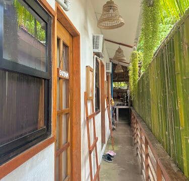 Narrow outdoor hallway in Siargao with wooden doors, bamboo fence, hanging lamps, and lush greenery on the right side.