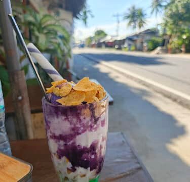 A tall glass of halo-halo dessert with cornflakes on top sits on an outdoor table along a lively street in Siargao.