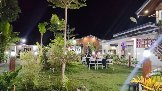 Outdoor dining area at night in Siargao, with tables, chairs, lush plants, and well-lit modern buildings in the background.