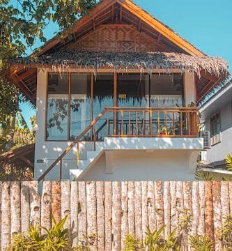 Two-story tropical Siargao house with a thatched roof, wooden fence, and potted plants in front, under a clear sky.