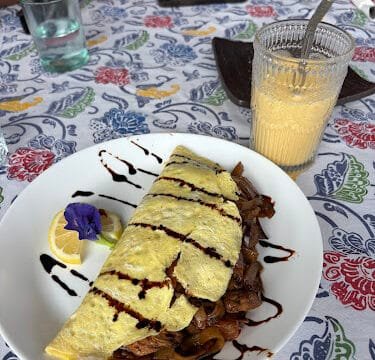 Omelette with vegetables and sauce, lemon slice, juice in a glass, and hands around a colorful Siargao tablecloth.