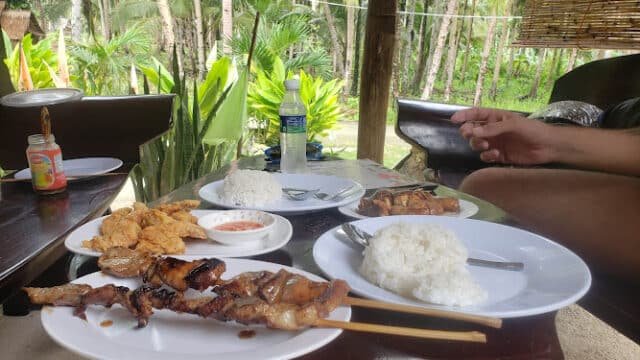 Grilled meat skewers, rice, and dipping sauce served outdoors in Siargao with palm trees in the background.