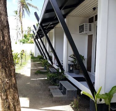 Small white modern cabins with black metal supports and lush plants, nestled under Siargao’s sunny sky with palm trees.