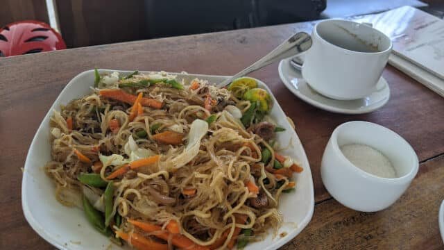 A plate of stir-fried noodles with vegetables, a cup of coffee, and a sugar bowl on a wooden table in Siargao.