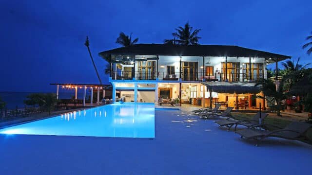 Modern Siargao beachfront villa at dusk with a glowing pool, lounge chairs, and palm trees under a deep blue sky.