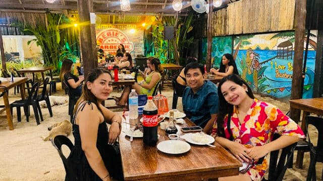 Three people smile at a wooden table in a lively, Siargao-inspired tropical restaurant with food and drinks.