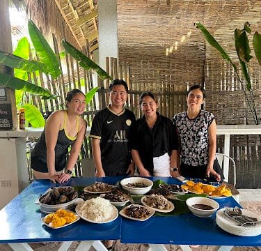 Four people smile behind a blue table filled with Filipino dishes, enjoying an open-air, tropical Siargao setting.