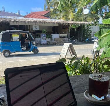 Tablet and iced coffee on a table, with a street, tuk-tuk, and Siargao’s palm trees visible outside the window.
