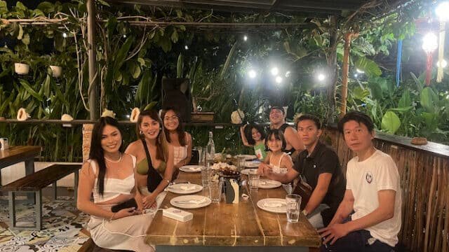 Eight people sit around a wooden table at a Siargao outdoor restaurant with lush greenery and string lights.