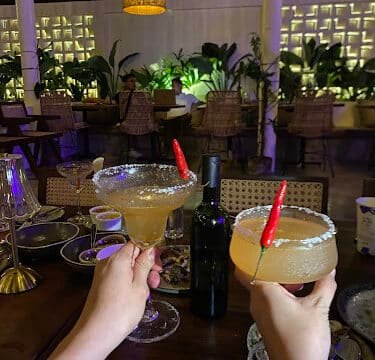 Two hands hold margarita glasses with chili garnish over a dinner table in a stylish, plant-filled Siargao restaurant.