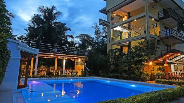 Illuminated outdoor swimming pool beside a multi-story building and patio at dusk in Siargao, surrounded by lush greenery.