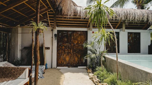 A tropical Siargao villa patio with wooden doors, palm trees, a lounge area, and a pool under a thatched roof.