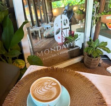 A cup of latte with latte art on a woven placemat at a cozy Siargao cafe, surrounded by plants and glass doors.