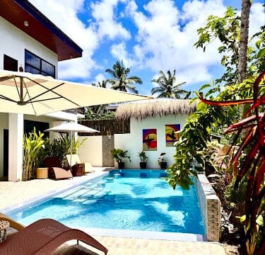 Sunny Siargao backyard with a pool, lounge chairs, umbrellas, tropical plants, and a modern house in the background.
