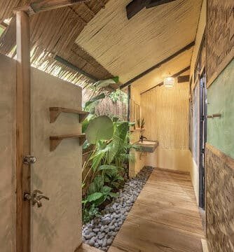 Outdoor rustic bathroom in Siargao with wooden floors, stone pathway, and lush green plants beneath a thatched roof.