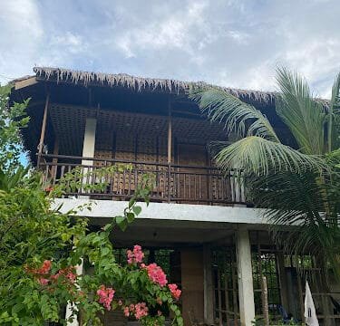 A charming two-story Siargao house with a thatched roof, wooden balcony, and lush green plants and flowers in front.