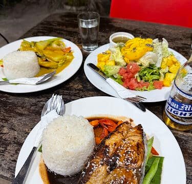 Three plates of food with rice, fish, salad, and curry on a rustic table in Siargao, with drinks and utensils.