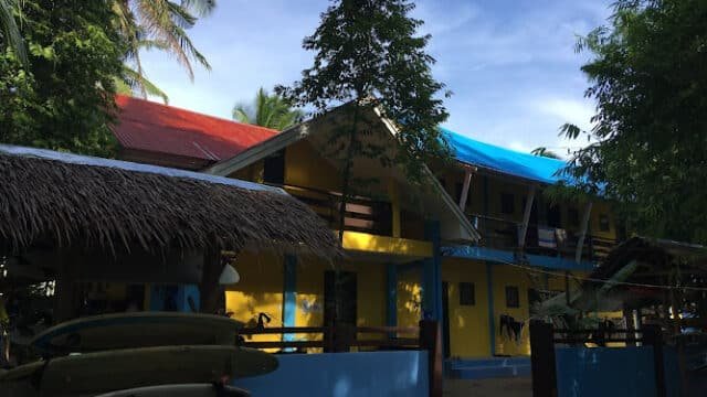 Two-story yellow building with red and blue roofs, surrounded by trees and surfboards in Siargao.