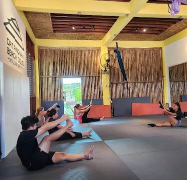 People in a gym class on Siargao sit on mats doing V-sit exercises, reaching for their toes with arms extended.