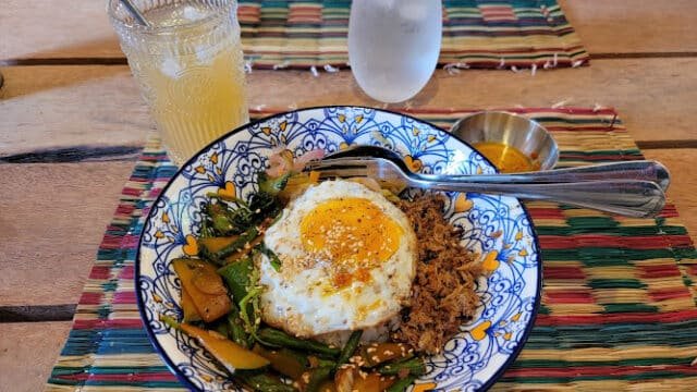 A bowl of rice with a fried egg, vegetables, and meat, served Siargao-style next to a drink and glass of water on the table.