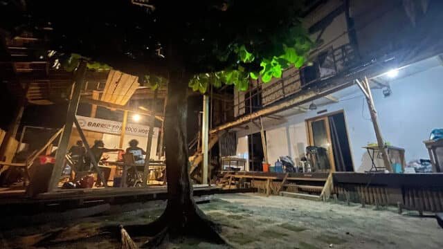 A tree stands in a sandy Siargao courtyard at night, with a lit wooden building and people gathered under a roof.