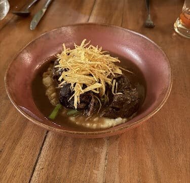 A bowl of braised meat on mashed potatoes, topped with crispy shoestring potatoes, served Siargao-style on a wooden table.