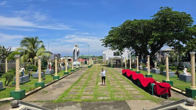 A person stands on a garden pathway in Siargao, surrounded by statues, columns, trees, and red-covered tables under a bright blue sky.