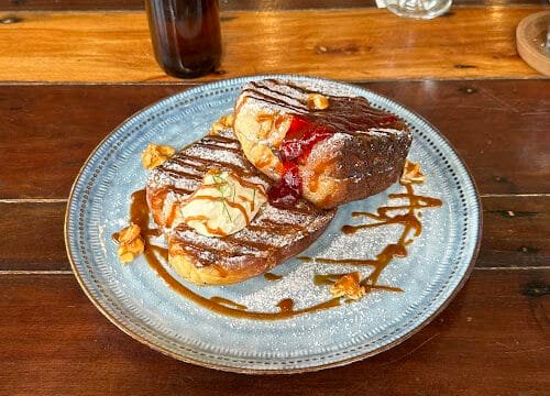 Two pieces of French toast topped with whipped cream, jam, nuts, and caramel drizzle served Siargao-style on a blue plate.