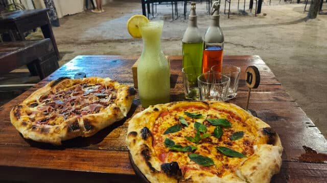 Two pizzas, a jug of lemonade, and condiments on a wooden table at a Siargao outdoor restaurant.