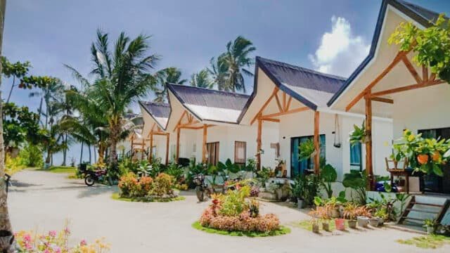 Row of white cottages with peaked roofs in Siargao, surrounded by palm trees and gardens on a sunny day.