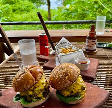 Two cheeseburgers on wooden boards with fries, soda, and sauce, set outdoors with a lush Siargao green backdrop.