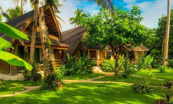 Thatched-roof cottages in Siargao, surrounded by lush greenery, palm trees, and a bright, sunny sky.