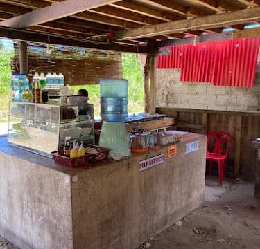 Outdoor food stall in Siargao with a display case, bottled water, condiments, and a red chair under a wooden roof.