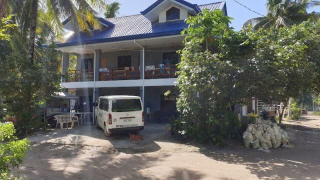 Two-story Siargao house with a blue roof, white van outside, trees, and bags piled by the entrance.