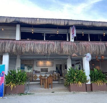 Outdoor view of a thatched-roof Siargao restaurant with wooden tables, lush plants, and open seating under a blue sky.