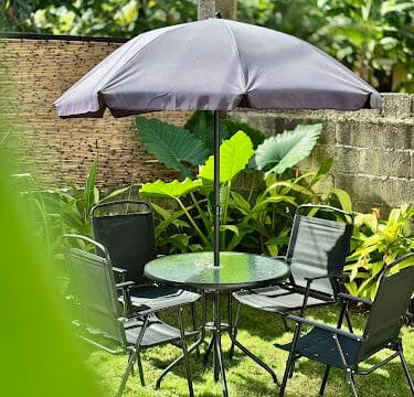 A round table with six black chairs and a large umbrella in a sunny Siargao garden with lush green plants.