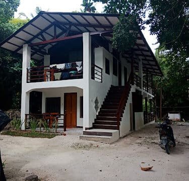 Two-story white house in Siargao with wooden stairs, balcony, and a motorcycle parked in front, surrounded by lush trees.