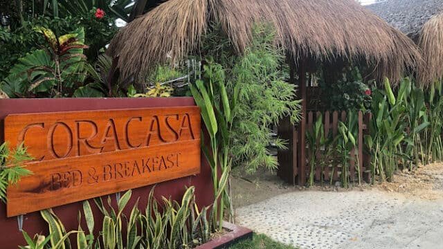 Wooden sign reading Coracasa Bed & Breakfast in front of a thatched-roof building with lush greenery in Siargao.