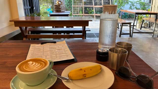 A cappuccino, banana, menu, and jam on a wooden table in a Siargao café with outdoor seating—plus water, cups, and sunglasses.