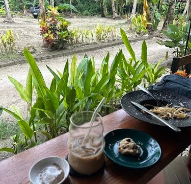 A wooden table with iced coffee, a pastry, pasta, and lush greenery and palm trees in the Siargao background.