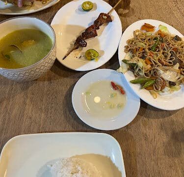 A Siargao-inspired table set with rice, soup, grilled meat skewer, noodles with vegetables, and a dipping sauce.