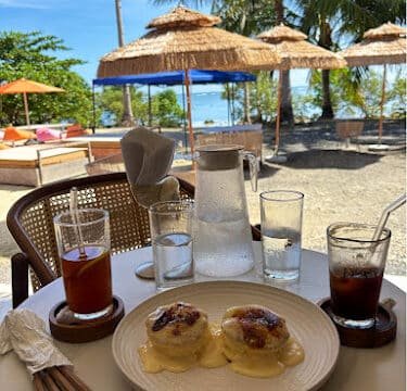 A table with drinks and pancakes overlooks Siargao's tropical beach, dotted with palm trees and sun umbrellas.