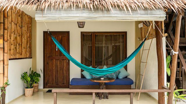 A turquoise hammock hangs on a porch with a bamboo roof in Siargao, near a wooden door, bench, and potted plants.