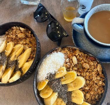 Two smoothie bowls with granola, banana, and chia seeds, served Siargao-style next to coffee, sunglasses, and a shot glass on a table.