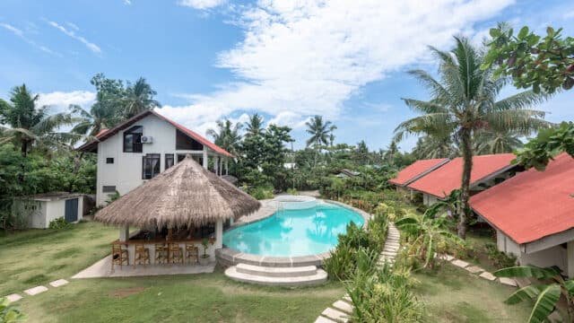 A tropical Siargao resort with a pool, palm trees, a thatched-roof bar, and white buildings under a blue sky.