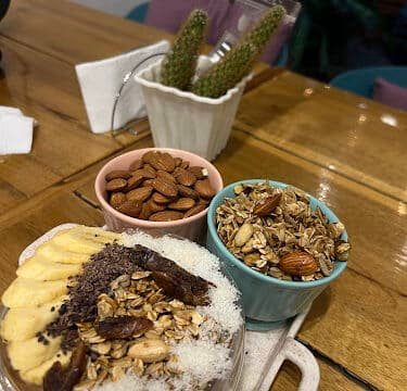 A bowl topped with banana, dates, coconut, and granola, inspired by Siargao, with side bowls of almonds and granola on a wooden table.