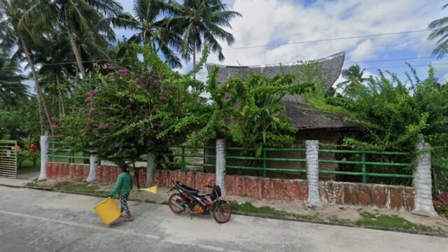 A person walks by a green fence and tropical plants in Siargao, with a parked red motorcycle on a sunny day.