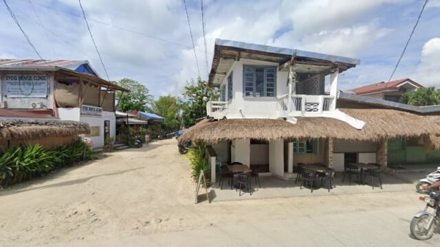 A white two-story building with a thatched roof and outdoor seating on a sunny day, reminiscent of Siargao village charm.