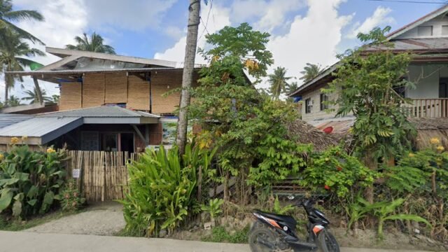 A black scooter parked on a street in Siargao, in front of tropical plants and houses with bamboo and wood walls.